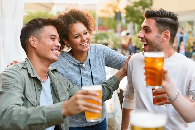 Eine Gruppe fröhlicher gemischtrassiger Freunde, die einander anschauen, während sie alkoholisches Bier auf der Terrasse eines Straßencafés in der Stadt trinken