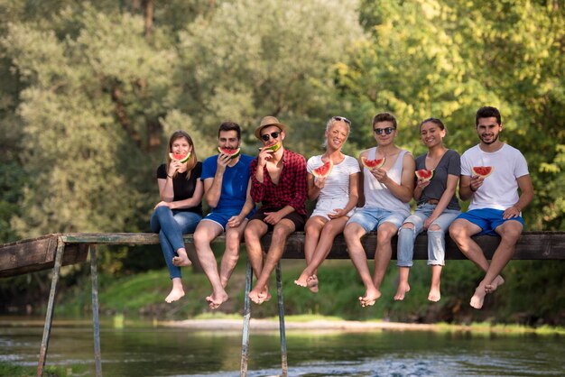 Eine Gruppe junger Freunde genießt Wassermelone, während sie auf der Holzbrücke über den Fluss in wunderschöner Natur sitzt