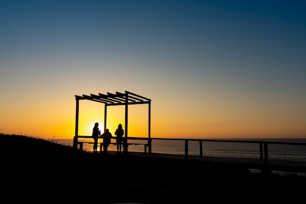 Eine Gruppe junger Leute in einem Aussichtspunkt, der den Sonnenuntergang am portugiesischen Strand von Sao Pedro de Moel - Portugal beobachtet.