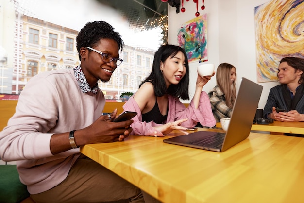 Eine Gruppe junger Leute, Mädchen und Jungs, arbeiten in einem Café aus der Ferne an Laptops, unterhalten sich, machen Selfies.