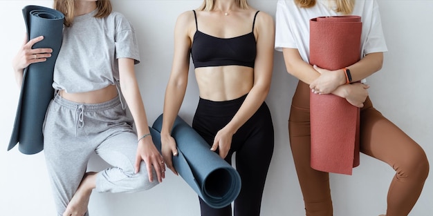 Eine Gruppe junger Sportmädchen mit Yogamatten, die gegen eine weiße Wand stehen Freundinnen im Fitnessstudio, die sich nach Fitness oder Yoga im Innenbereich entspannen