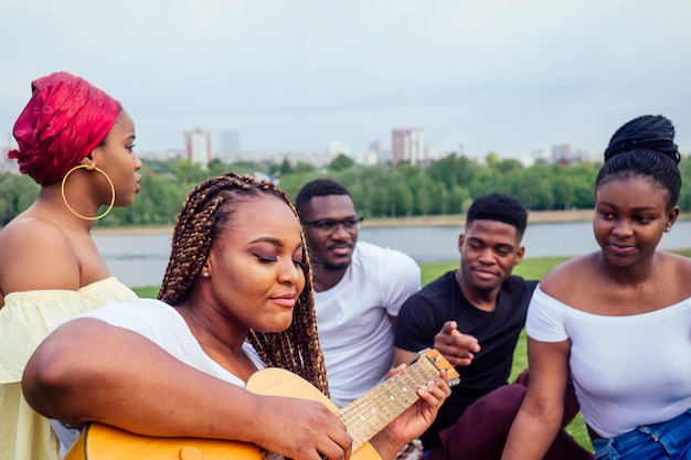 Eine Gruppe lächelnder, glücklicher afroamerikanischer Freunde, die in der Kampagne im Freien Gitarre spielen, picknicken