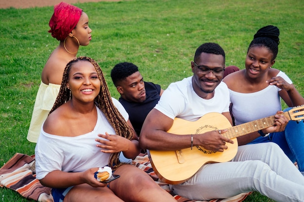 Eine Gruppe lächelnder, glücklicher afroamerikanischer Freunde, die in der Kampagne im Freien Gitarre spielen, picknicken