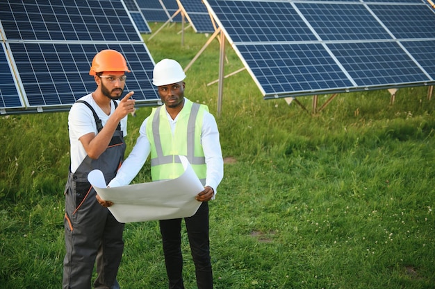 Eine Gruppe mehrethnischer Menschen in Sicherheitshelmen steht in einer Solarstation, zwei Ingenieure und Techniker untersuchen den Plan der Paneele im Freien
