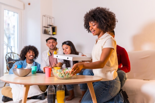 Eine Gruppe multiethnischer Freunde auf einem Sofa, die Pizza essen und alkoholfreie Getränke auf einer Hausparty trinken. Der junge Mann kommt mit der Pizza von der Straße