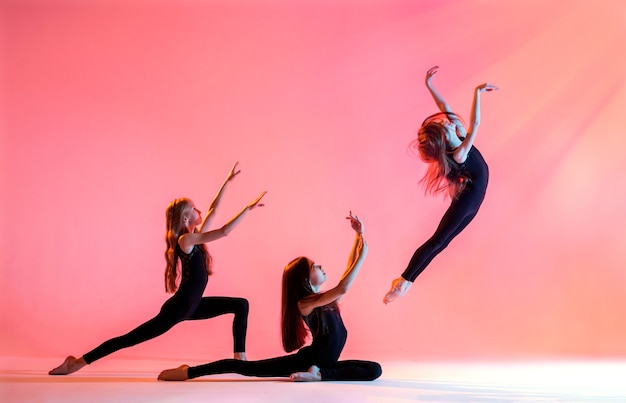 Eine Gruppe von drei Ballettmädchen mit langen fließenden Haaren in schwarzen, eng anliegenden Anzügen tanzt auf einem roten Hintergrund