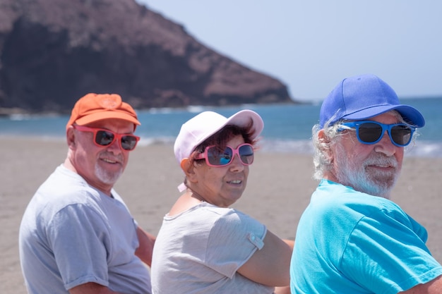 Eine Gruppe von drei reifen Kaukasiern, die mit Mützen und Sonnenbrillen am Strand sitzen, drehen sich um, um auf die Kamera Horizon über dem Wasserkopierraum zu schauen