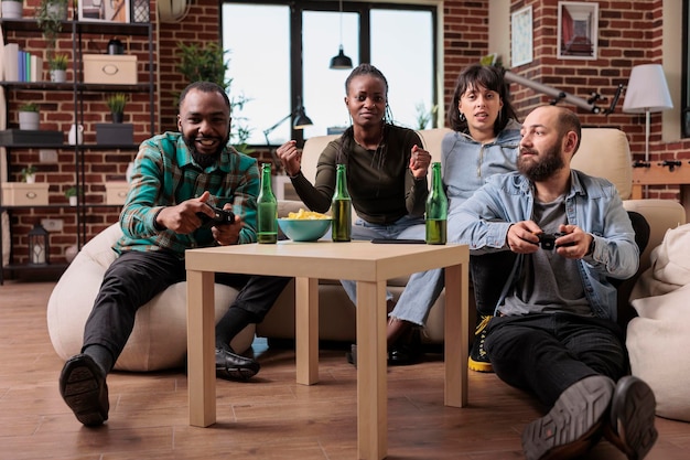 Eine Gruppe von Freunden, die sich mit Videospielwettbewerben amüsieren und Bier bei einem geselligen Beisammensein trinken. Wettbewerbsfähige Leute spielen auf der Fernsehkonsole und genießen Freizeitaktivitäten und Unterhaltung.