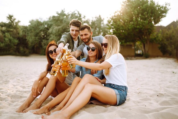 Eine Gruppe von Freunden jubelt und trinkt Bier am Strand, junge Freunde entspannen sich und machen ein Picknick im Sommer.