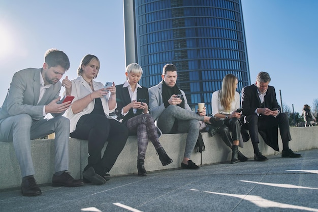 Eine Gruppe von Geschäftsleuten schaut in ihre Smartphones, während sie im Freien sitzen Pausenzeit Warten auf ein Meeting