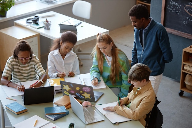 Eine Gruppe von Kindern arbeitet an einem großen Tisch im Unterricht, während ein männlicher Lehrer zuschaut