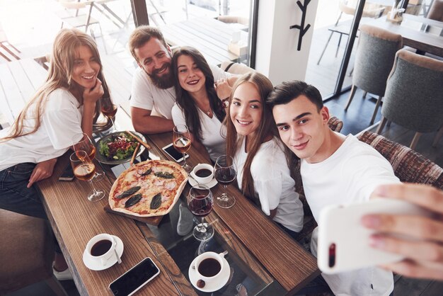 Eine Gruppe von Menschen macht ein Selfie-Foto in einem Café. Die besten Freunde versammelten sich an einem Esstisch, aßen Pizza und sangen verschiedene Getränke