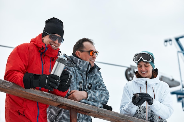 Eine Gruppe von Skifahrerfreunden auf dem Berg ruht sich aus und trinkt Kaffee aus einer Thermoskanne im Hintergrund des Skilifts.