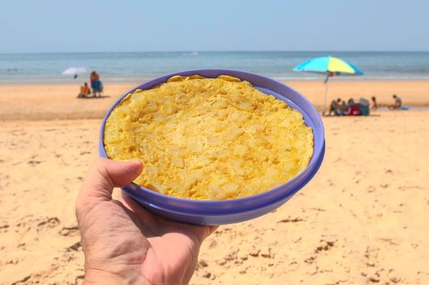 Eine Hand, die ein Kartoffelomelett in einer Tupperware hält. Ein Strand im Hintergrund.