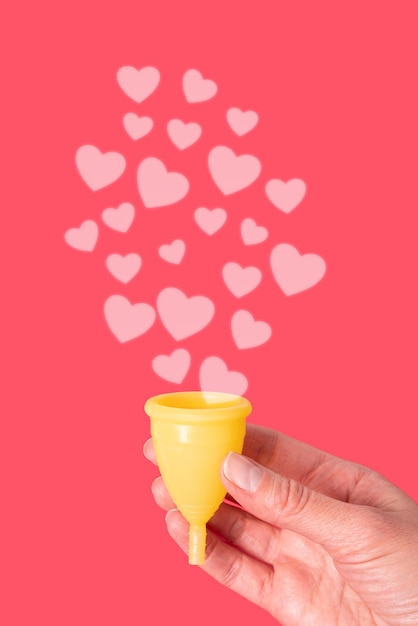 Eine Hand, die eine gelbe Menstruationstasse auf rotem Hintergrund mit rosa Herzen als Menstruationstropfen hält Frauengesundheitskonzept Umweltfreundliches Konzept Null-Abfall-Konzept