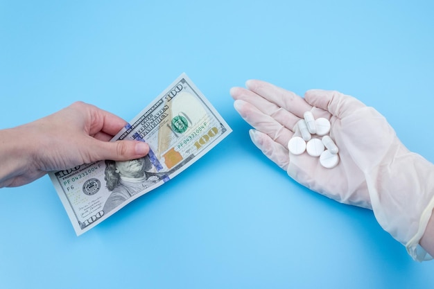 Eine Hand gibt hundert Dollar für eine Hand in einem medizinischen Handschuh mit einer Pille auf blauem Hintergrund