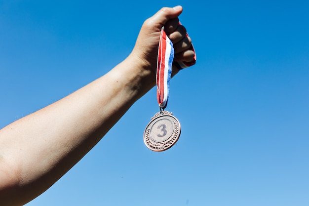 Eine Hand hält eine Bronzemedaille auf dem dritten Platz mit dem Himmel im Hintergrund. Siegeskonzept