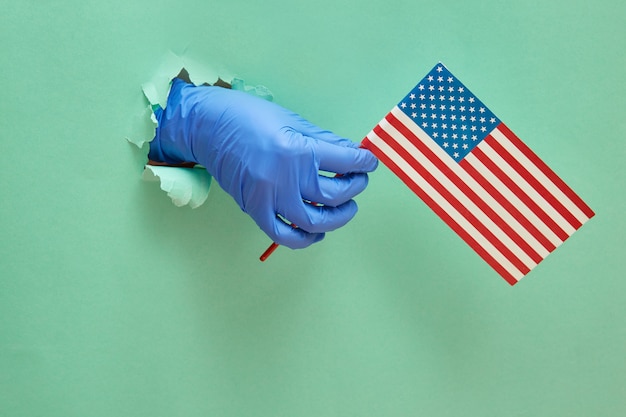 Eine Hand in einem blauen Nitrilschutzhandschuh hält die amerikanische Flagge. Der moderne Trend ist ein Loch im Papier mit Kopierraum.