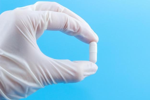 Eine Hand in einem weißen medizinischen Handschuh hält eine Kapsel in Nahaufnahme auf blauem Hintergrund. Das Konzept der Gesundheitsversorgung oder medizinischen Versorgung.