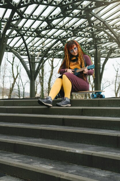 Eine helle Frau sitzt auf einem Longboard auf der Straße und spielt Gitarre und Ukulele auf der Straße