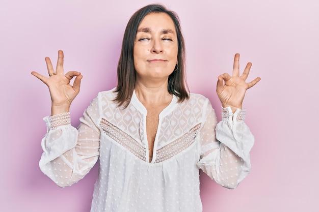 Eine hispanische Frau mittleren Alters in legerer Kleidung entspannt sich und lächelt mit geschlossenen Augen und macht eine Meditationsgeste mit den Fingern beim Yoga-Konzept