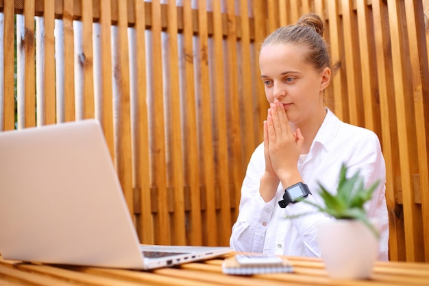 Eine hoffnungsvolle Frau in weißem Hemd sitzt in einem Straßencafé und arbeitet am Laptop, wartet auf wichtige E-Mails und betet für viel Glück bei der Online-Unterrichtsarbeit