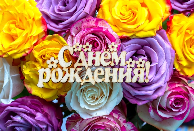 Eine Holzplatte für einen Blumenstrauß mit der Aufschrift Happy Birthday auf Russisch, die auf einem hellen, schönen Rosenstrauß liegt