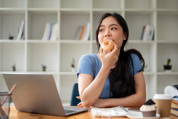 Eine hübsche asiatische Frau genießt ihren Snack und isst Donuts an ihrem Schreibtisch zu Hause