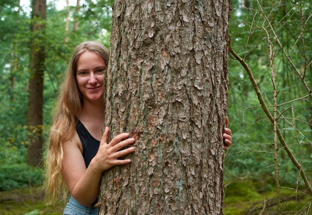 Eine hübsche blonde kaukasische Frau mit langen Haaren lächelt und umarmt einen Baumstamm im Wald