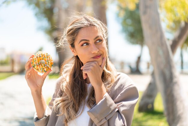 Eine hübsche blonde uruguayische Frau hält einen Donut im Freien und denkt über eine Idee nach und schaut nach.