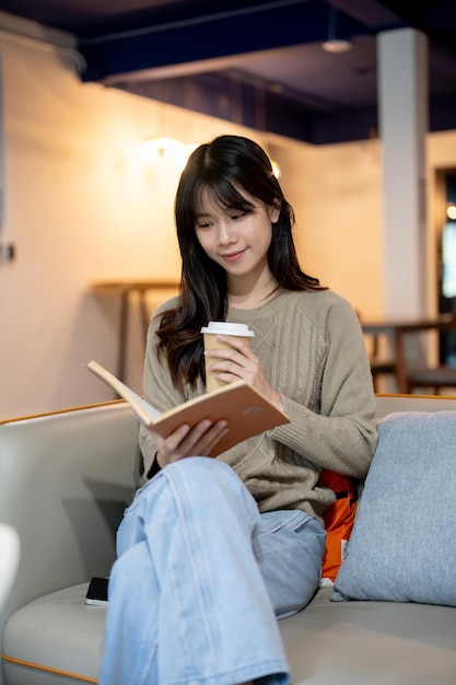 Eine hübsche, glückliche asiatische Frau liest ein Buch und trinkt Kaffee, während sie in einem gemütlichen Café sitzt