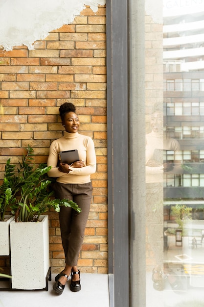 Eine hübsche junge afroamerikanische Geschäftsfrau mit einem digitalen Tablet steht an der Backsteinmauer im Büro im industriellen Stil