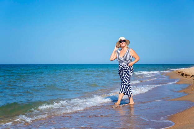 Eine hübsche lächelnde ältere Frau in einem Kleid spaziert am Sandstrand entlang. Urlaubsreisen und aktiver Lebensstil