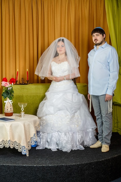 Eine jüdische Braut mit verhülltem Badekengesicht und ein Bräutigam in einer Synagoge stehen während einer Zeremonie vor Chupa. Vertikales Foto
