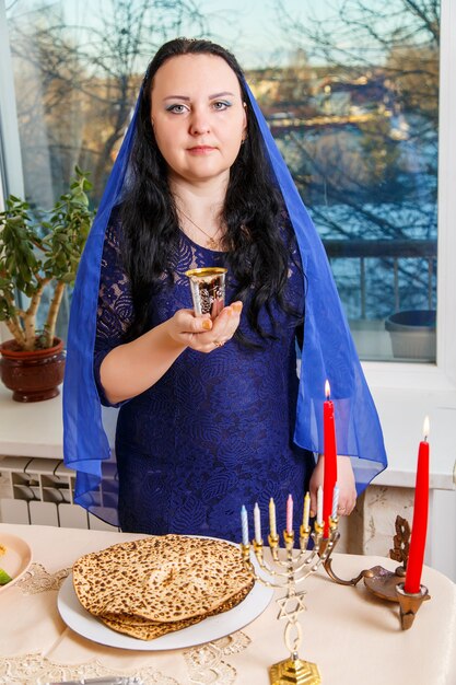 Eine jüdische Frau, deren Kopf mit einem blauen Umhang am Pessach-Seder-Tisch bedeckt ist, macht Kiddusch für Wein. Vertikales Foto