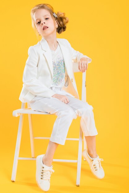 Eine Jugendliche in der weißen Kleidung sitzt auf einem Stuhl auf einer gelben Wand.