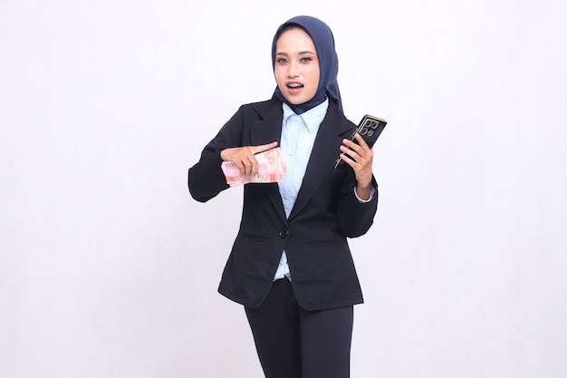 Eine junge asiatische Bürofrau, die einen Hijab trägt, steht schockiert da und trägt und zeigt auf ein Smartphone-Gadget
