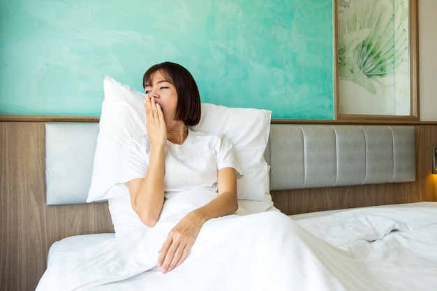 Eine junge asiatische Frau in einem weißen T-Shirt sitzt und gähnt mit der Hand über den Mund auf einem Bett mit Morgenlicht Aktivitäten beim Aufwachen