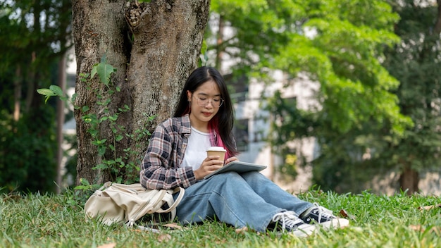 Foto eine junge asiatische frau sitzt unter einem baum in einem campuspark mit einer kaffeetasse und einem tablet