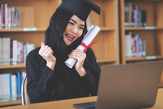 Eine junge asiatische Hochschulabsolventin drückt Freude und Aufregung aus, um ihren Abschluss vor einem Laptop zu feiern, der einen Remote-Videoanruf mit ihren Eltern zu Hause macht