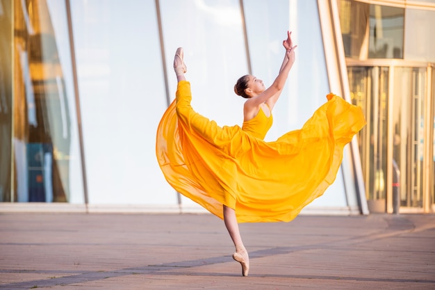 Eine junge Ballerina in einem langen, fliegenden gelben Kleid tanzt vor der Kulisse eines Stadtbildes