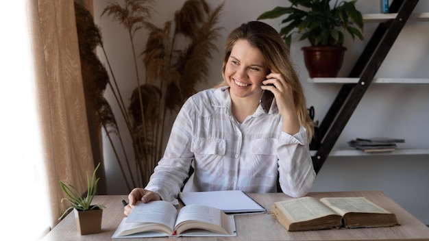 Eine junge beratende Ärztin spricht am Telefon, konsultiert und sitzt an einem Holztisch mit Büchern und einem Notizblock Persönlicher Berater