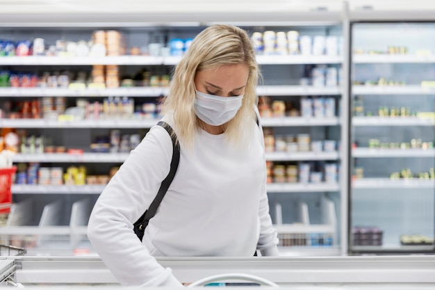 Eine junge blonde Frau in einer medizinischen Maske wählt Tiefkühlkost in einem großen Supermarkt aus. Gesunde Ernährung und Vegetarismus. Coronavirus Pandemie.