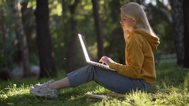 Eine junge blonde Frau in Freizeitkleidung mit einem Laptop sitzt auf dem grünen Gras. Die Sonne beleuchtet den Laptop. Bücher liegen in der Nähe. Studieren in der Natur. 4K UHD