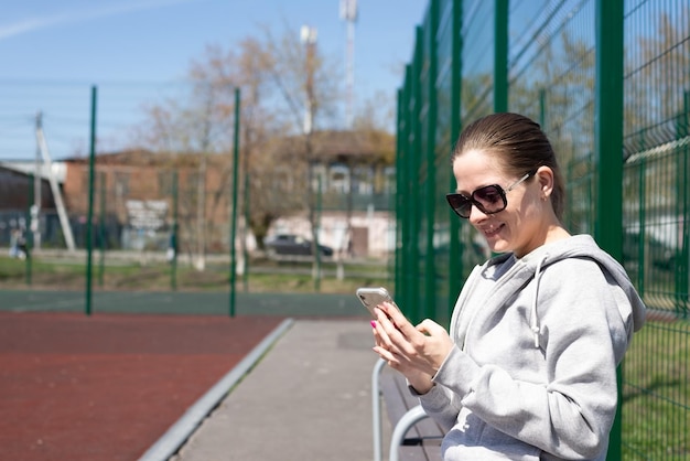 Eine junge blonde Frau in Sportbekleidung ruht sich aus, nachdem sie auf einer Bank im Stadion Sport gelaufen ist und in sozialen Netzwerken kommuniziert