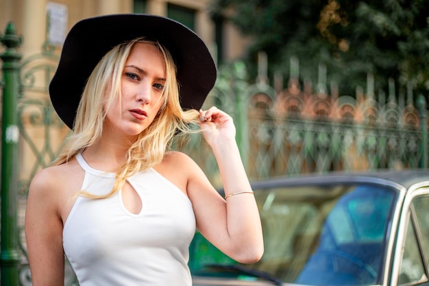 Eine junge blonde Frau posiert auf einem Auto auf der Straße, ein weibliches Modemodell mit einem eleganten Outfit und einem Auto