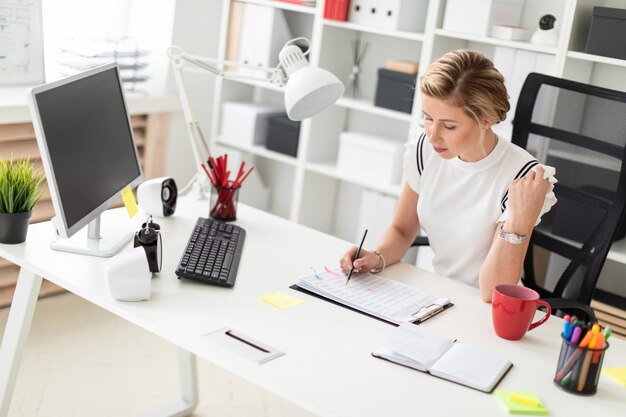 Eine junge blonde Frau sitzt an einem Computertisch im Büro und schreibt einen Bleistift in das Dokument.