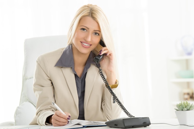 Eine junge blonde Geschäftsfrau, die im Büro telefoniert und mit einem Lächeln auf ihrem Gesicht in die Kamera schaut.