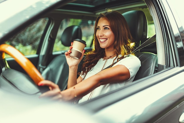 Eine junge erfolgreiche lächelnde Frau hält eine Tasse Kaffee in der Hand, während sie ihr neues Auto fährt.