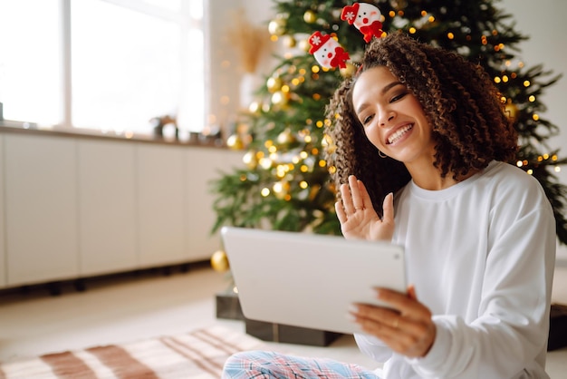 Eine junge Frau auf dem Hintergrund eines Weihnachtsbaums mit Geschenken mit einem Tablet hat einen Videoanruf oder Video-Chat.